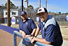 The baseball and softball coaches for Louisburg College, two men, lean across a blue fence and smile.