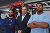 Hurricane football team head coach stands in the middle of two of his players who are team captains in the weight room.