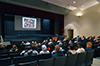 Audience members seated in an audience listen as a speaker describes an image on the screen 