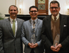 Four men standing side by side smiling. The two men in the middle are holding alumni awards. 